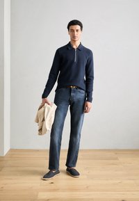 Marine zip-krage genser, blå jeans, grå slip-on sko og beige jakke holdt i hånden; glatt tekstur, casual passform og nøytrale toner.