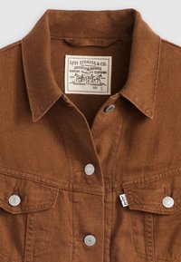Camisa de ganga castanha com colarinho, dois bolsos no peito, botões de pressão prateados e uma etiqueta com a inscrição "Levi Strauss & Co., tamanho S."