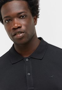 Czarna koszulka polo z teksturowanej tkaniny, z trzema guzikami na plastrze i małym haftowanym logo po lewej stronie.