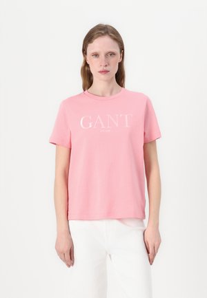 Růžové bavlněné tričko s krátkými rukávy, kulatým výstřihem a bílým textem "GANT" přes hruď. Hladká textura, uvolněný střih.