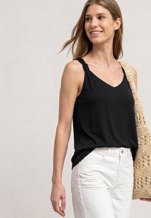 Jeune femme portant un débardeur noir et un pantalon blanc, portant un sac à bandoulière beige en tricot, souriante et regardant sur le côté.