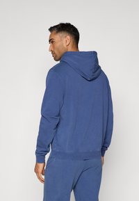 Kék pamut kapucnis pulóver, húzózsinórral, bordás mandzsettákkal és alsó szegéllyel. Puha tapintású, laza szabású, látható varrási részletekkel.