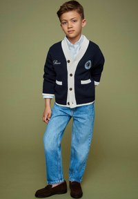 Námornícky sveter s krémovými akcentmi, gombíkmi a logom; svetlomodrá košeľa; denimové džínsy; hnedé topánky. Neformálny, vrstvený outfit pre deti.