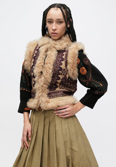 Γιλέκο από συνθετική γούνα με πολύπλοκα μοτίβα σε μωβ και μπεζ αποχρώσεις· διαθέτει μαύρα μανίκια με κεντήματα λουλουδιών. Συνδυάζεται με πλισέ φούστα σε ελιά πράσινο.