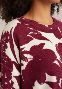 Adīts džemperis ar apaļu izgriezumu. Iezīmējas tumši sarkanu ziedu rakstu uz krēmkrāsas fona, mīksta tekstūra un riboti piedurkņu gali.