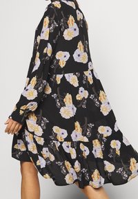 Zwarte jurk met lange mouwen en een vloeiende gelaagde rok, met een bloemenpatroon in witte, gele en oranje tinten.