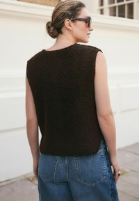 Bruine, textuurvolle mouwloze top met een ronde halslijn, gecombineerd met blauwe high-waisted denim jeans. Zijkantweergave met detail van schouder en rug.