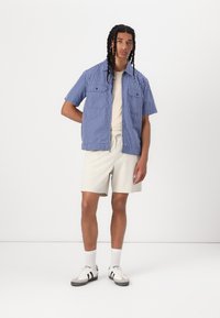 Kurzärmliges blau-weiß kariertes Shirt mit zwei Brusttaschen, kombiniert mit beigen Shorts und grau-weißen Sneakers. Einfarbiger Hintergrund.