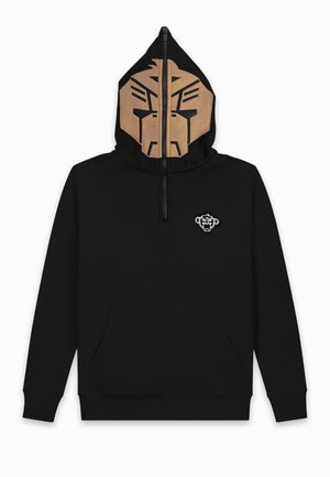 Person trägt einen schwarzen Kapuzenpullover mit Halb-Reißverschluss-Gesichtsmaske, die ein abstraktes beiges Gesicht zeigt, hellblaue zerrissene Jeans und beige Sneakers.