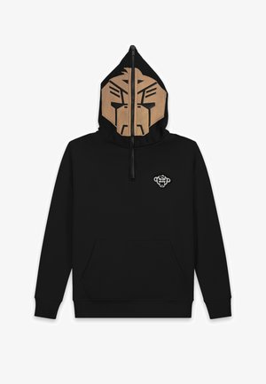 Person trägt einen schwarzen Kapuzenpullover mit Halb-Reißverschluss-Gesichtsmaske, die ein abstraktes beiges Gesicht zeigt, hellblaue zerrissene Jeans und beige Sneakers.