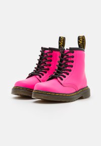 Botines de cuero rosa con cordones negros, costuras amarillas, suela negra y una lengüeta amarilla con la marca "AirWair".