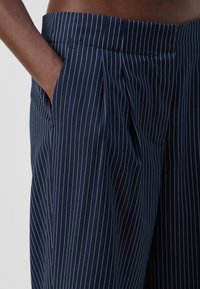 Pantalons bleu marine à rayures fines avec une taille plate, des poches latérales et une ceinture ajustée. Tissu texturé avec des lignes verticales blanches.