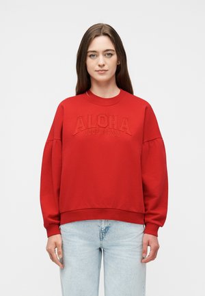 Mujer joven con sudadera roja con la inscripción "ALOHA JOOP! JEANS" en relieve en el frente y vaqueros azul claro, posando contra un fondo liso.