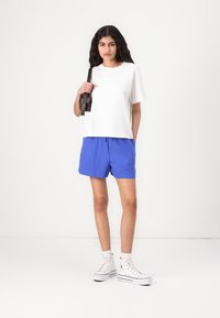 Hvit t-skjorte med korte ermer kombinert med blå shorts med snøring, komplementert av hvite høye sneakers og en mørk skulderveske.