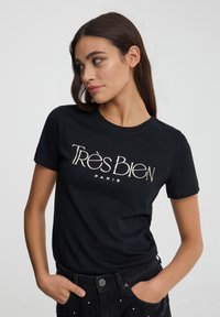 T-shirt nera in cotone con maniche corte e scollo tondo, caratterizzata da testo metallici "Très Bien" e "PARIS" sulla parte anteriore.