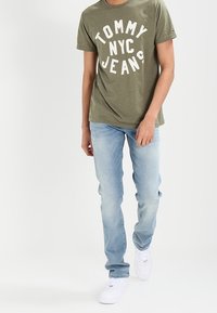 olivově zelené bavlněné tričko s nápisem "TOMMY NYC JEANS" v bílé barvě, v kombinaci se světle modrými džíny a bílými teniskami.