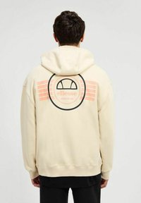 Sudadera crema con un gran gráfico circular negro y rosa en la parte posterior, que presenta el nombre de la marca "ellesse" y rayas horizontales.