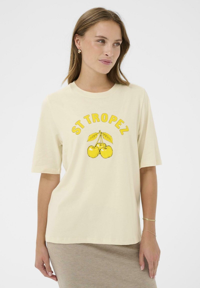 Femme portant un t-shirt oversized crème avec des cerises jaunes et le texte "ST TROPEZ", associé à une jupe marron clair et des bracelets en or.