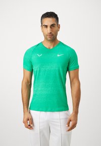 Zelena sportska majica kratkih rukava s V-izrezom, s teksturiranom površinom i bijelim logotipima Nike i emblemom marke.