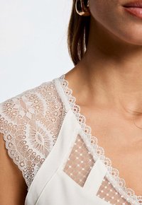 Top blanc avec des épaules en dentelle à bord festonné et des détails à pois, mettant en avant une texture et des éléments de design complexes.