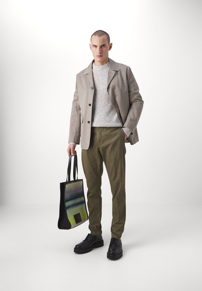 Lichtgrijze jas, gebreide textuurtrui, olijfgroene broek, zwarte schoenen en een zwarte tote bag met abstracte patroon, alles in een minimalistische setting.