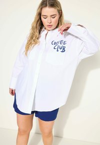 Camisa blanca de botones con estampado azul "Coffee Club" en el bolsillo, con mangas largas y un corte relajado, combinada con pantalones cortos azul marino.