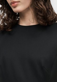 Γυναίκα με καστανά σγουρά μαλλιά που φοράει απλό μαύρο μπλουζάκι με στρογγυλή λαιμόκοψη, κομμένο ώστε να φαίνονται οι ώμοι, ο λαιμός και τα χείλη.