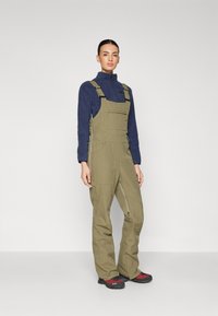 Olivgröna hängselbyxor med justerbara remmar, framfickor och dragkedjor vid anklarna, parat med en marinblå fleecepullover och röda skor.