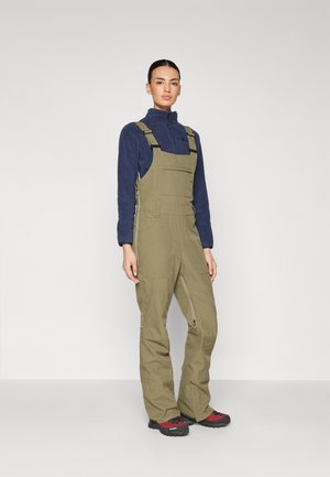 Olijfgroene bretels met verstelbare banden, voorzakken en ritsen bij de enkels, gecombineerd met een marineblauw fleece pullover en rode schoenen.
