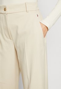 Cream-colored pants with a tailored fit, featuring a front pocket, button closure, and subtle branding details on the side.