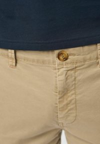 Gros plan d’un pantalon beige avec un bouton marron attaché à la taille, associé à une chemise bleu marine foncé rentrée.