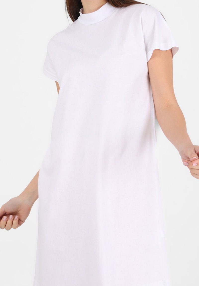 Femme portant une robe blanche unie à manches courtes avec un col rond haut sur fond blanc.