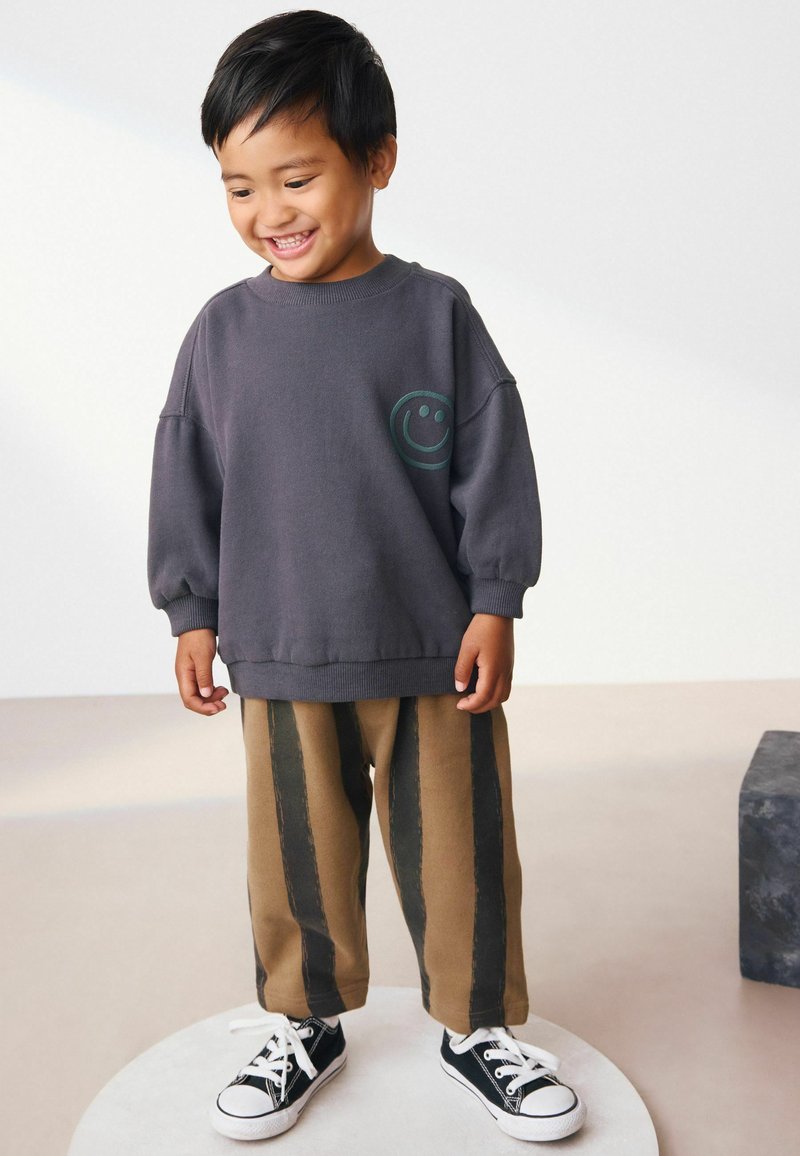 Bambino sorridente che indossa un felpa grigia con una faccina sorridente e dei pantaloni larghi a righe marroni e nere, in piedi su una piattaforma bianca rotonda.