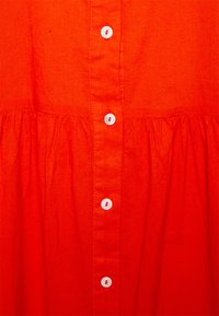 Vestido de algodão vermelho com cintura franzida, uma fila de botões brancos na frente e um tecido liso, ligeiramente texturizado.