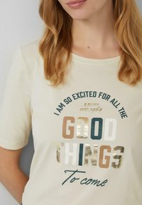 T-shirt beige clair en coton avec des manches courtes. Présente un texte imprimé en vert, or et marron. Encolure arrondie, coupe décontractée.