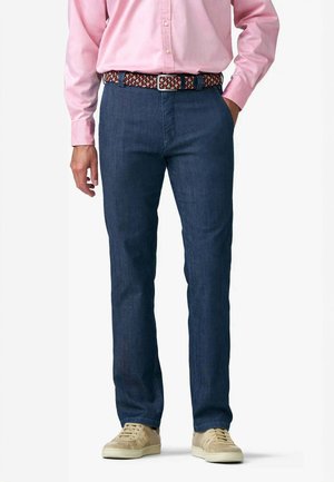 Uomo che indossa jeans blu scuro, sneakers beige, una camicia rosa con bottoni e una cintura intrecciata rossa, bianca e blu navy, una mano in tasca.