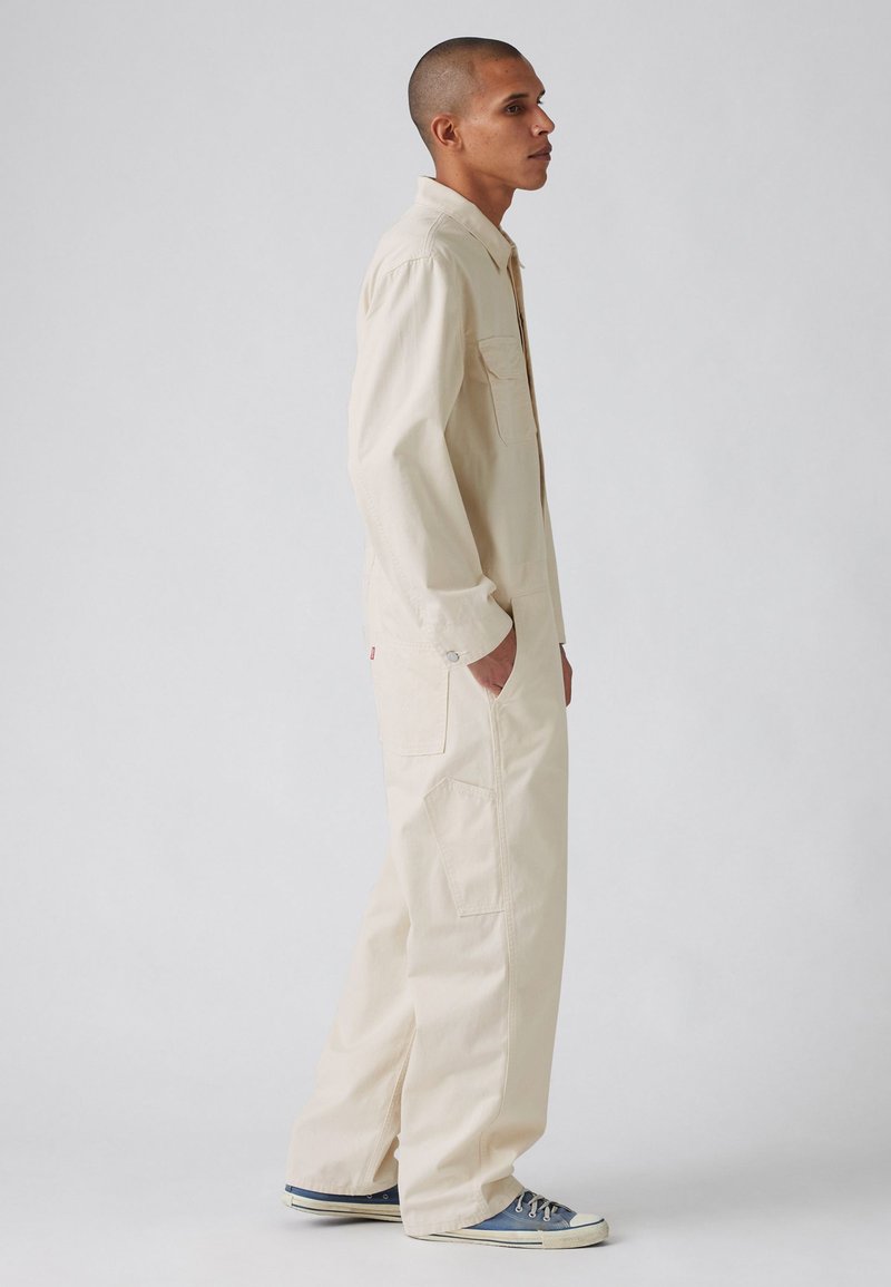 Combinaison légère beige clair avec un col, des manches longues et des poches avant ; coupe décontractée avec des jambes droites, assortie à des baskets bleues.