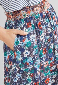 Main dans la poche d'une jupe plissée à motifs floraux avec des fleurs bleues, vertes, rouges et blanches, et une ceinture élastique brodée orange, associée à une chemise rayée.