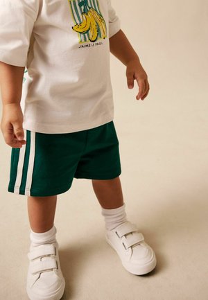 Kleinkind trägt weiße Klett-Sneaker, weiße Socken, grüne Shorts mit weißen Streifen und ein weißes T-Shirt mit einem Bananenmotiv und Text.