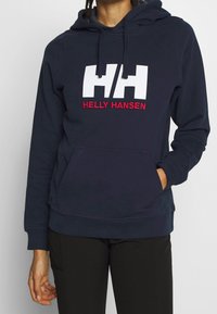 Mörkblå hoodie med framficka, prydd med vit "HH" logotyp och röd "HELLY HANSEN" text. Tillverkad av bomull med en mjuk känsla och justerbar huva.