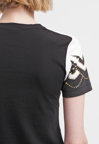 Černé tričko s krátkými bílými rukávy, které má zlatý geometrický vzor podél ramenního švu. Měkká látka a uvolněný střih.