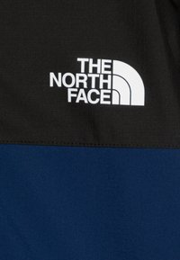 Črna in modra jakna z izrazito belim logotipom "The North Face" na črnem delu, izdelana iz gladkega, strukturnega materiala.