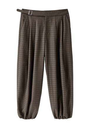 Femme posant avec un pull court beige, un débardeur blanc, un pantalon large marron à carreaux avec ceinture, et des talons hauts blancs pointus sur fond rose.
