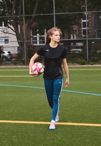 Black athletic shirt with Puma logo, blue sweatpants with white stripes, holding a red-and-white soccer ball, walking on green turf field.