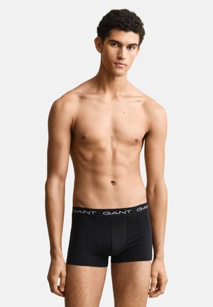 Jeune homme aux cheveux courts et bouclés, debout, face à l'avant, portant un boxer noir GANT sur un fond clair uni.