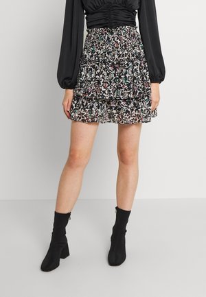 Vrouw draagt een zwarte gerimpelde top, een bloemenmouw mini-rokken met laagjes en zwarte enkellaarzen, staand tegen een eenvoudige lichte achtergrond.