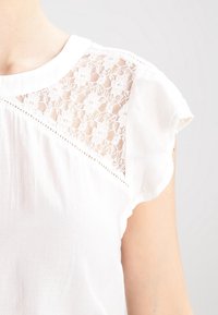 Blusa blanca con cuello redondo, panel de encaje transparente en el hombro con patrón floral y bordes de manga con volantes. Tejido suave y texturizado.