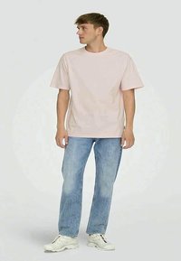 T-shirt di cotone rosa chiaro con maniche corte, abbinata a jeans di denim blu chiaro. Il modello si trova su uno sfondo semplice. Sneakers bianche presenti.
