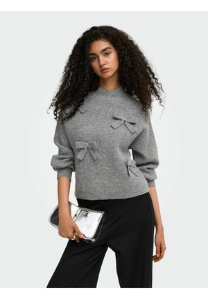 Femme aux longs cheveux bouclés porte un pull gris avec des nœuds en tissu et un pantalon noir, tenant une pochette argentée brillante.