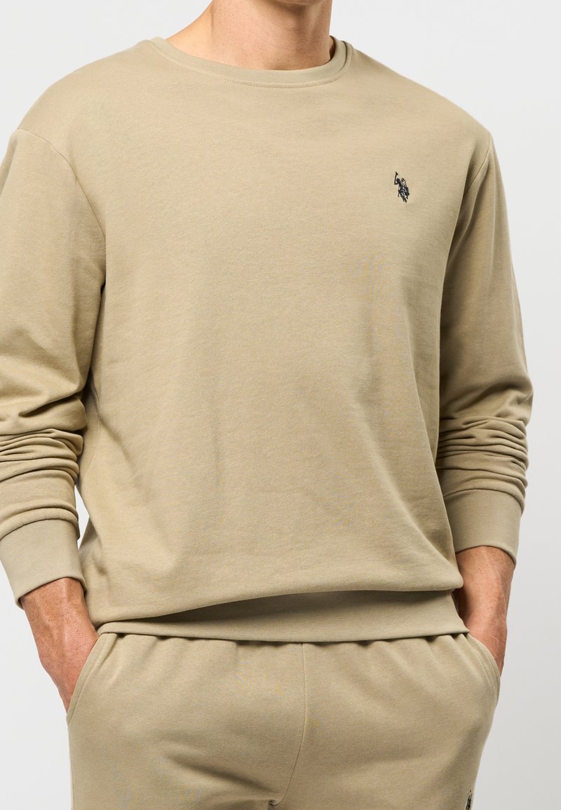 Homme portant un sweat-shirt beige à manches longues et un pantalon assorti avec un petit logo noir sur la poitrine gauche et les mains dans les poches.